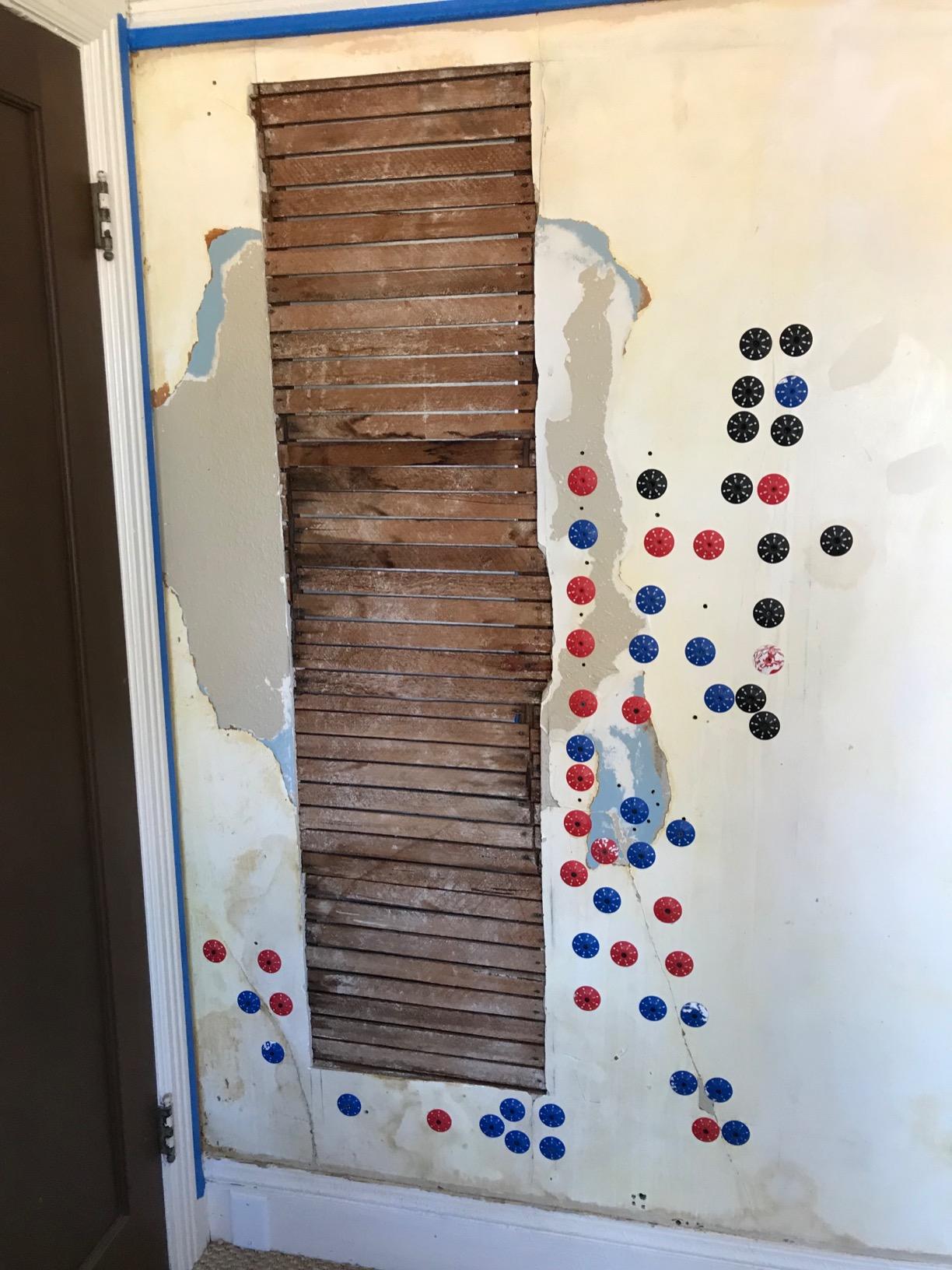 Damaged plaster wall exposing wooden lath, marked with red and blue adhesive injection points for reattachment
