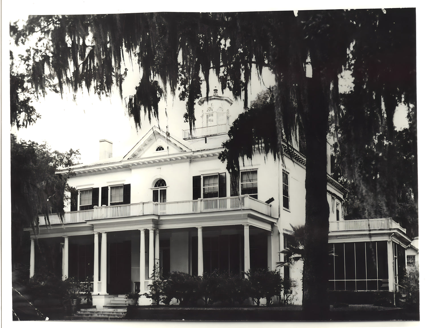 Black and white photograph of a large house with a prominent porch and trees in the foreground.