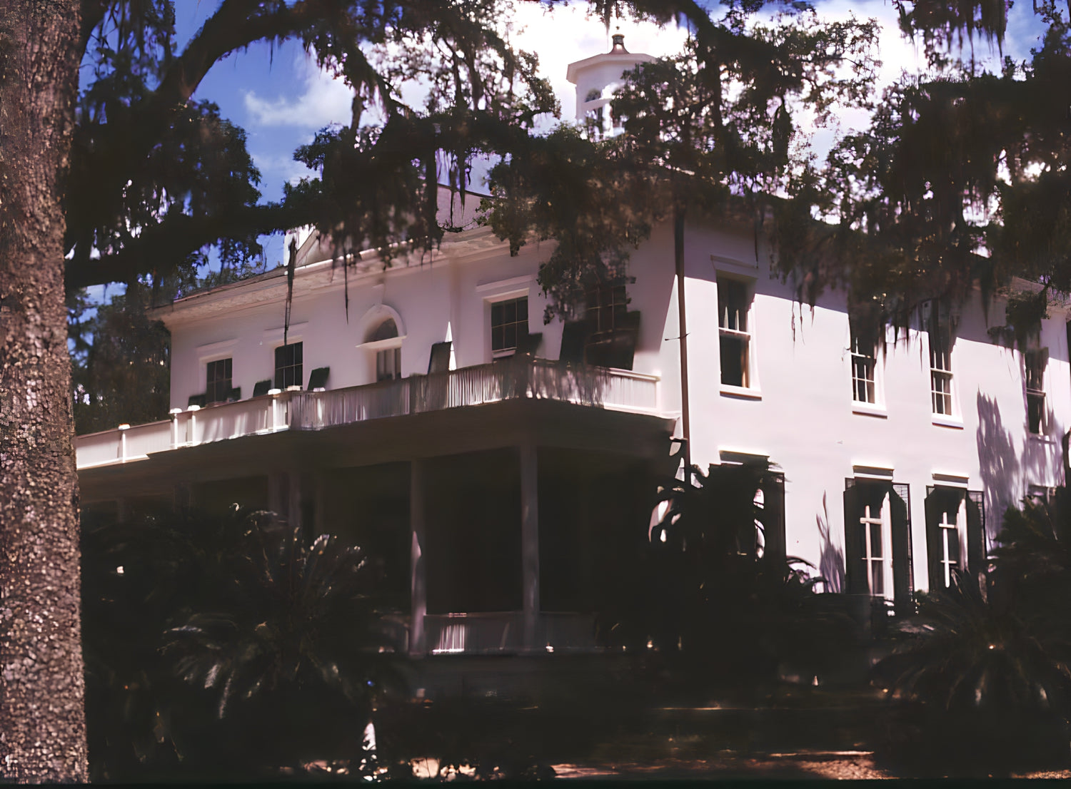 Large white mansion with columns and trees in front, under a blue sky.