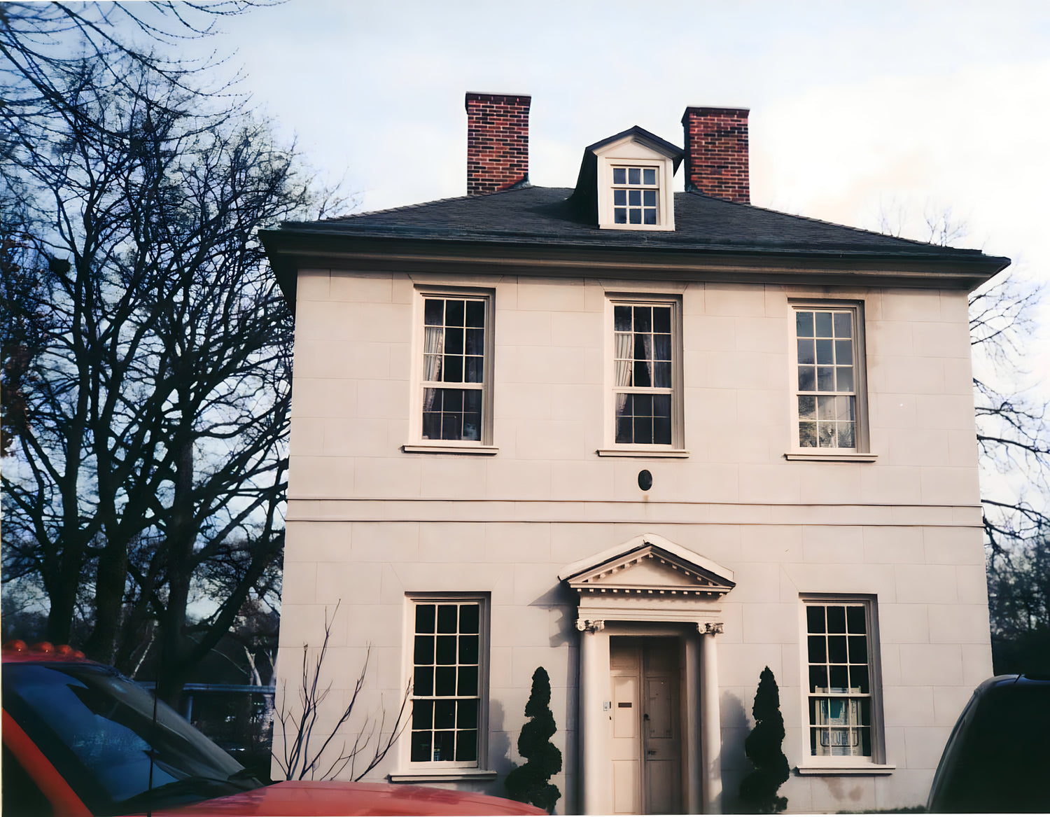 Two-story house with a central entrance and two chimneys, surrounded by trees.