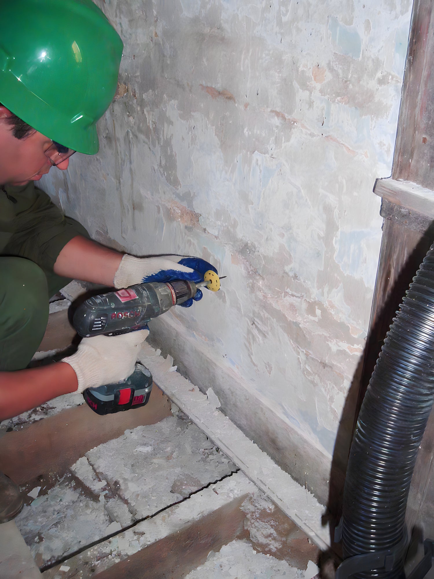Person using a drill on a concrete wall with a green helmet and gloves.