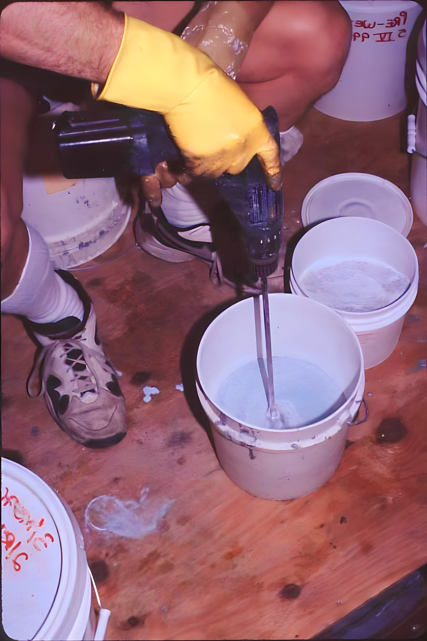 Person using a drill to mix contents in a bucket on a wooden floor with buckets around.