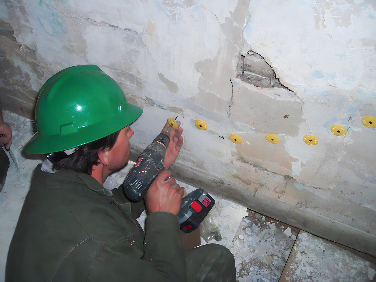 Person in a green hard hat using a power drill on a concrete wall.