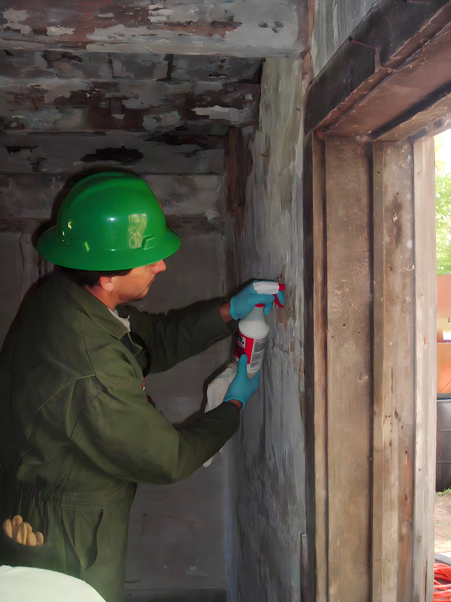 Person in green hard hat and coveralls applying a substance to a wall with a spray bottle.