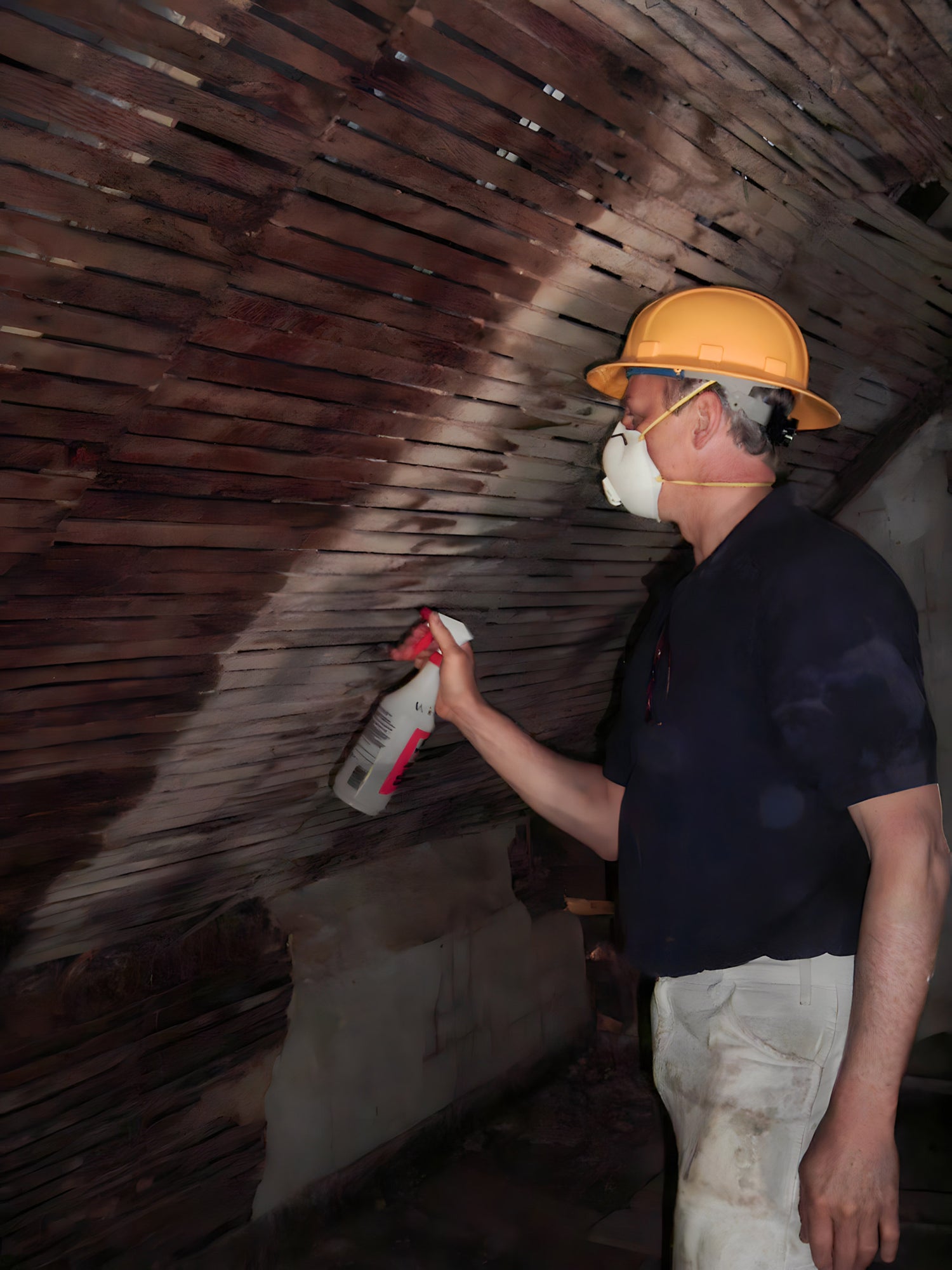 Person wearing a hard hat and protective mask, using a spray tool on a wooden ceiling.