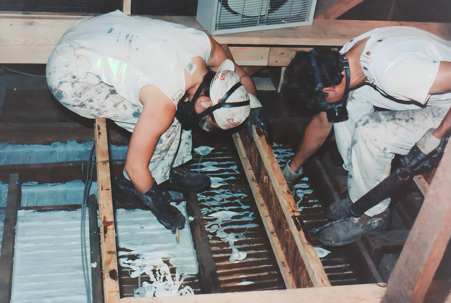 Two workers applying a white substance to wooden panels in a controlled environment.